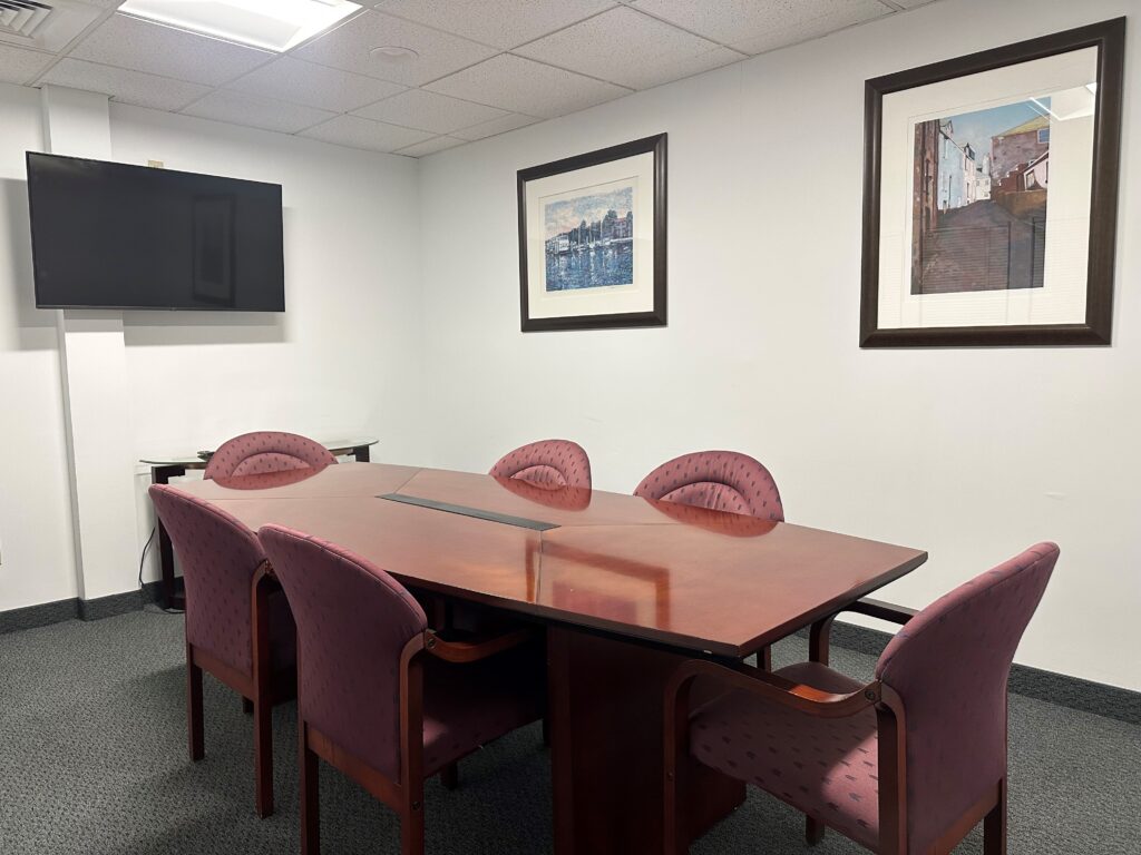Conference room with a polished wooden meeting table, upholstered chairs, wall-mounted television, and framed artwork on white walls, creating a professional setting.