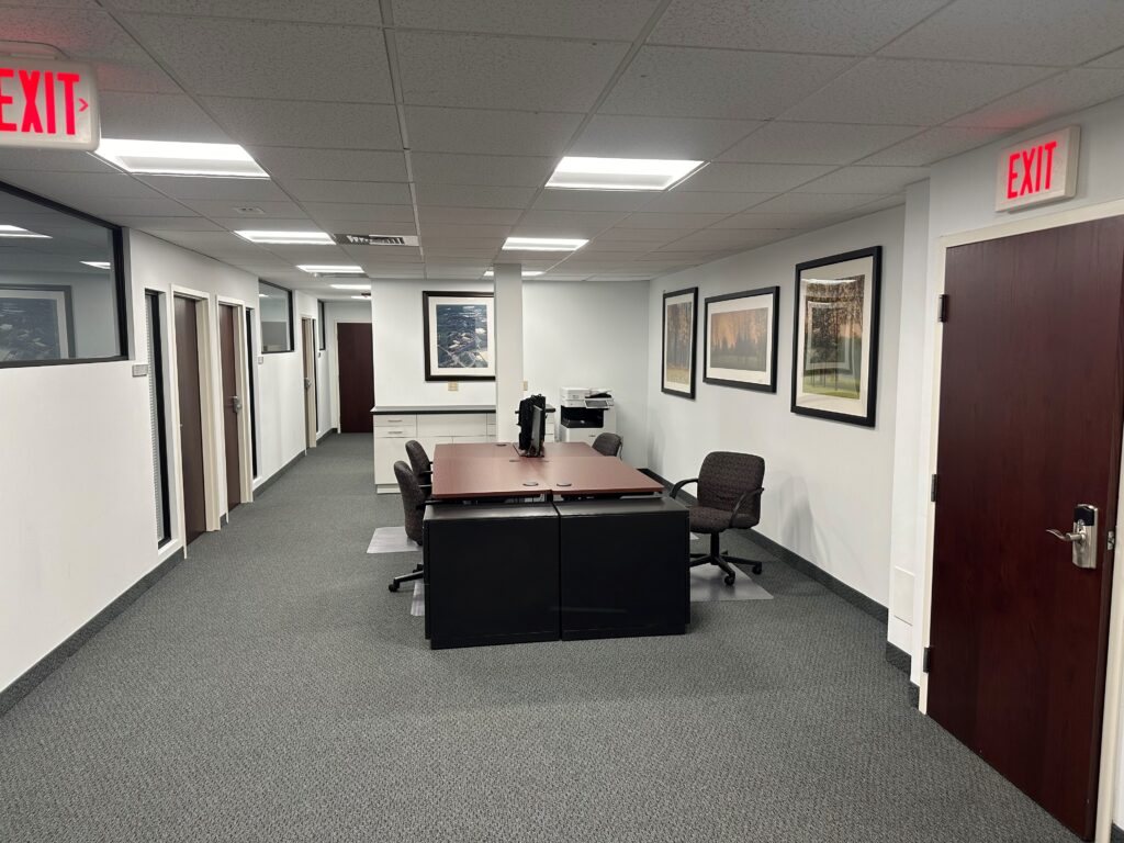 Professional office hallway with framed artwork on the walls, private offices with glass windows, a central meeting table, ergonomic chairs, and bright overhead lighting.
