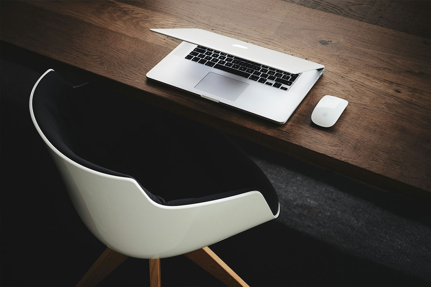 Modern workspace with a sleek white chair, wooden desk, partially open laptop, and wireless mouse, creating a minimalist and professional setup.